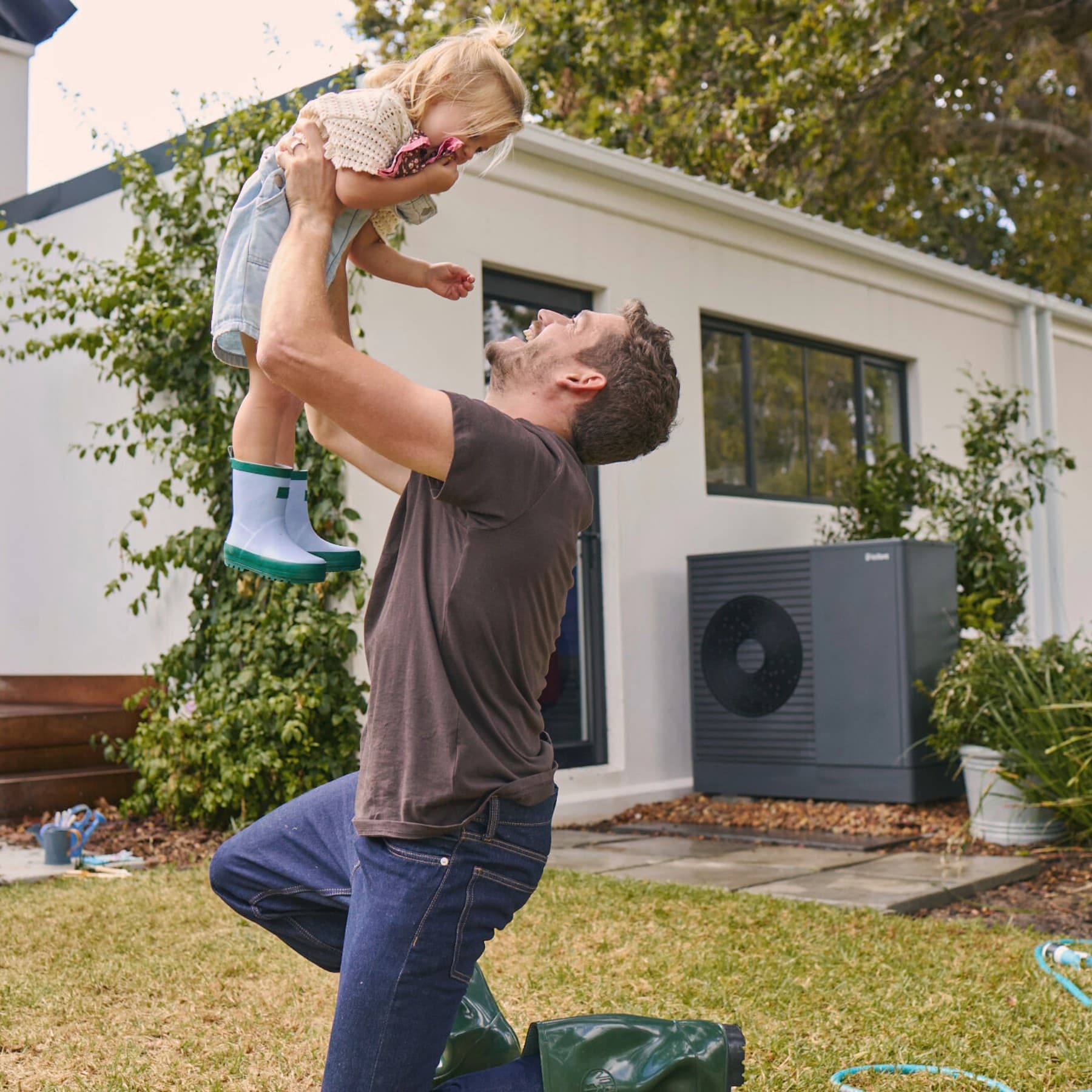 Padre che solleva la figlia in giardino con unità esterna della pompa di calore Vaillant aroTHERM plus