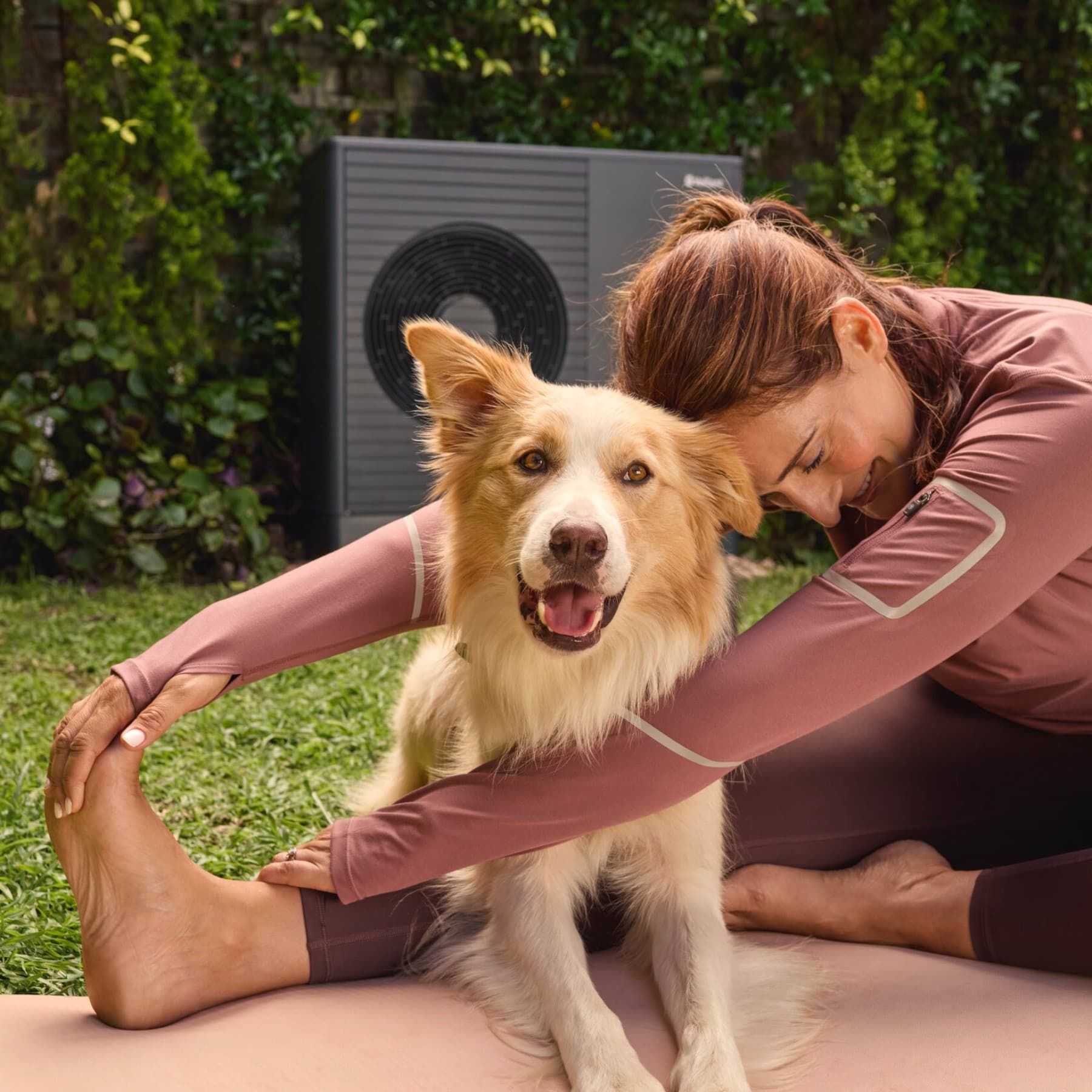 Donna che fa yoga in giardino con il proprio cane e pompa di calore Vaillant aroTHERM plus sullo sfondo