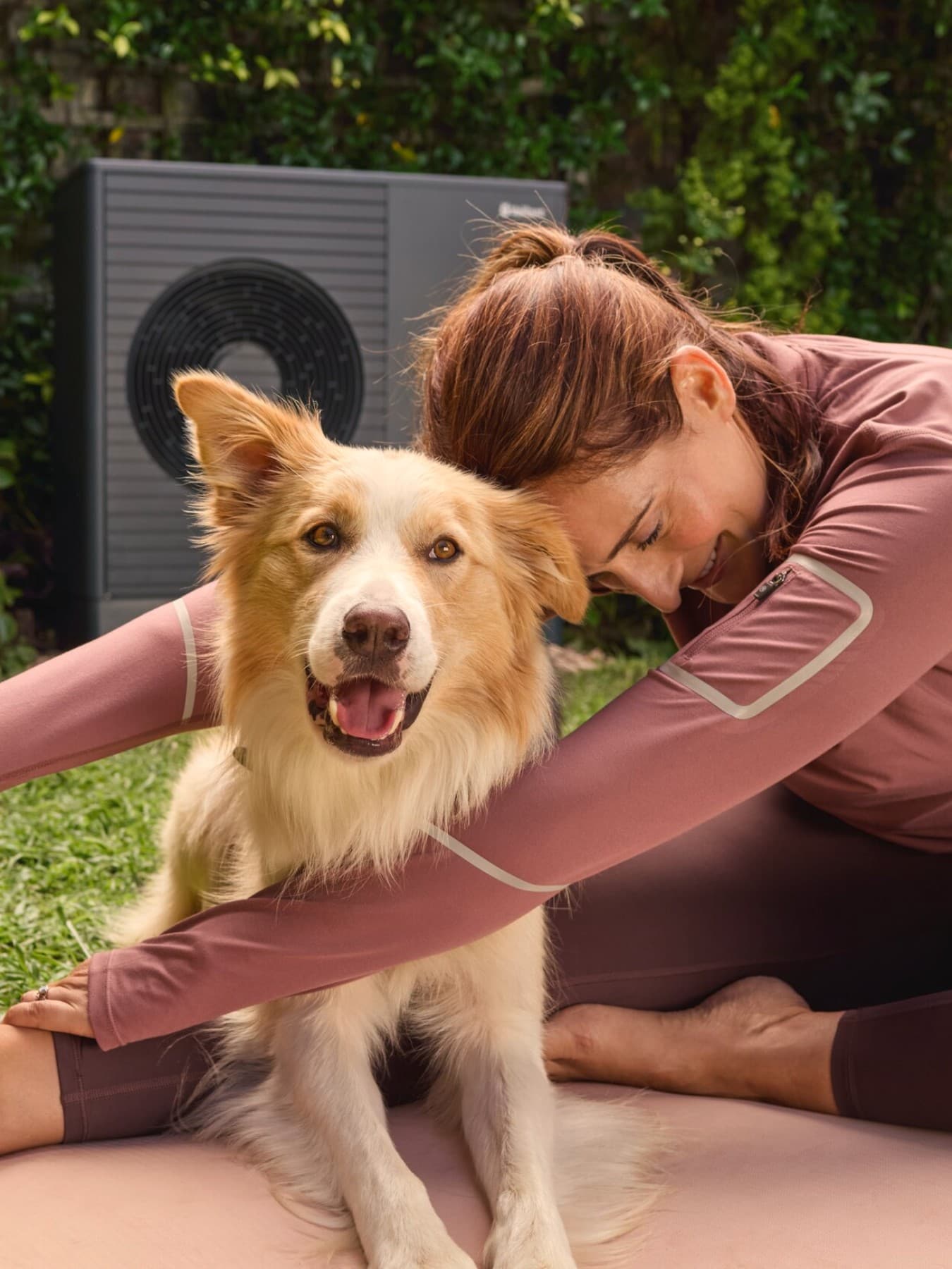 Donna che fa yoga in giardino con il proprio cane e pompa di calore Vaillant aroTHERM plus sullo sfondo