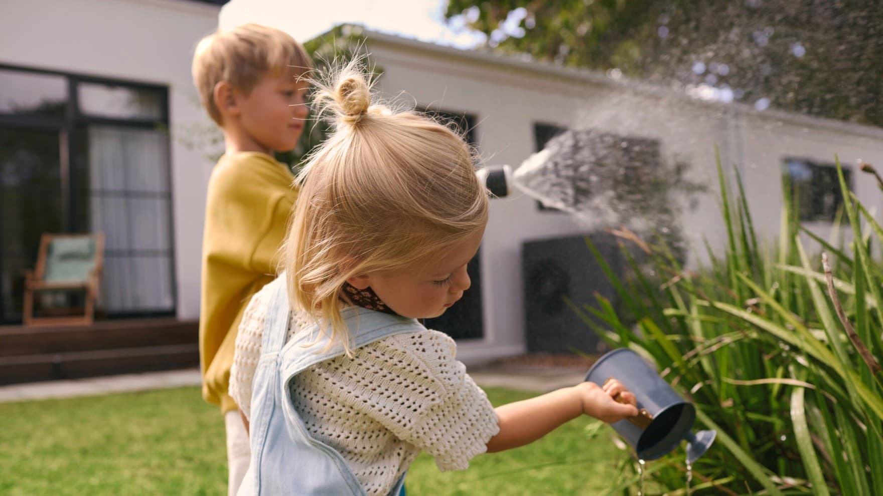 Bambini che annaffiano le piante in un giardino con una pompa di calore sullo sfondo.