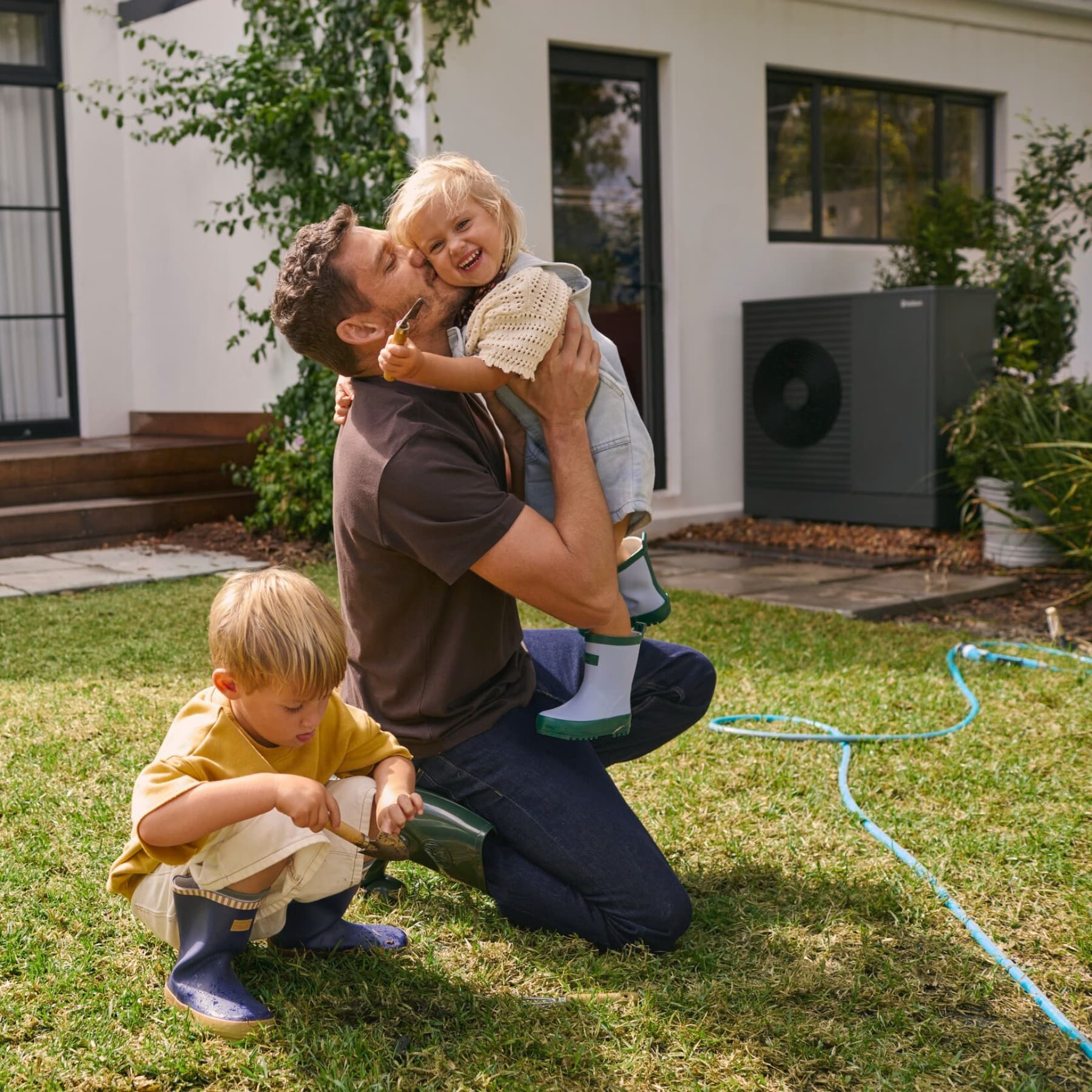 Padre che gioca con i figli in giardino con unità esterna della pompa di calore Vaillant aroTherm plus sullo sfondo